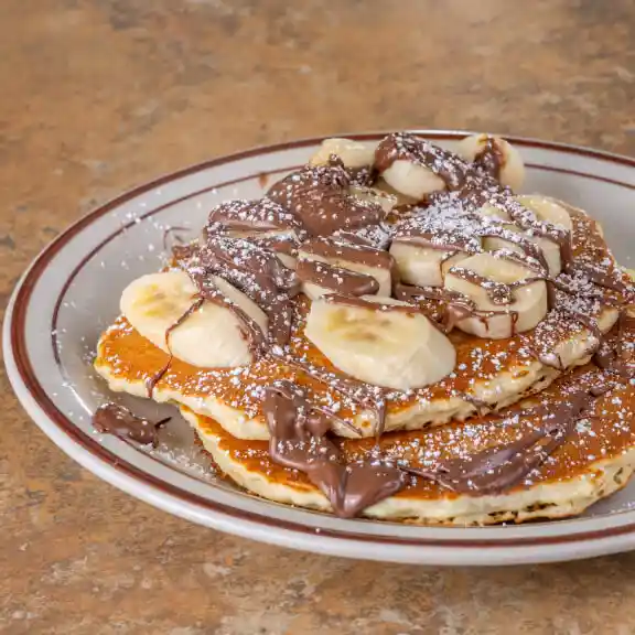 Stack of pancakes topped with bananas, chocolate drizzle, and powdered sugar